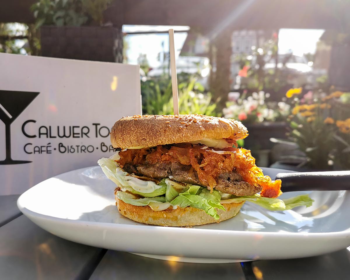 BBQ-Burger Burger mit Fleisch, Kraut und Salat auf einem weißen Teller. Speisekarte vom Calwer Tor sowie Blumen im Hintergrund.